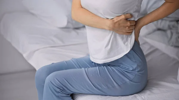 Partial view of woman sitting on bed in pajamas and suffering from abdominal pain — Stock Photo