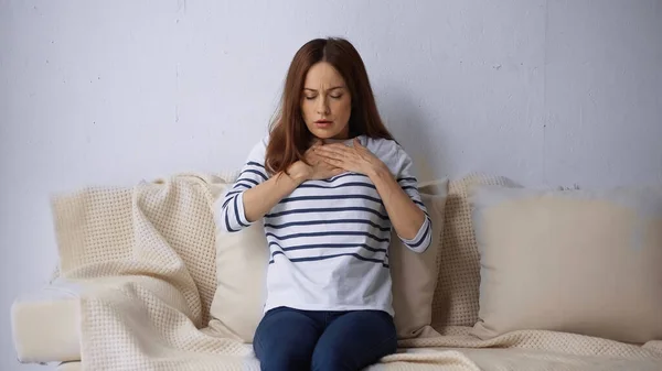 Femme avec les yeux fermés assis sur le canapé et ressentant une douleur dans la poitrine — Photo de stock