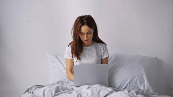 Brunette femme assise sur le lit à la maison et travaillant sur ordinateur portable — Photo de stock