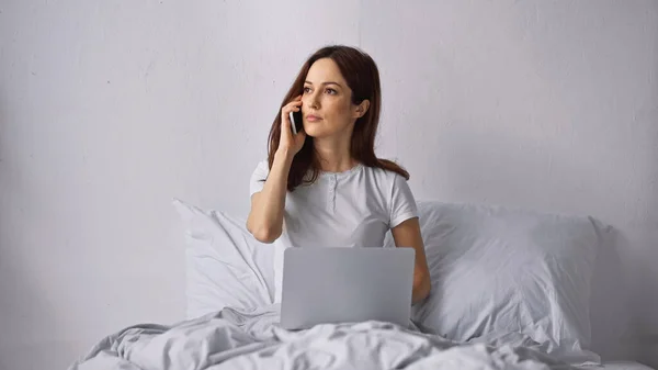 Брюнетка женщина сидит на кровати с ноутбуком и разговаривает по мобильному телефону — стоковое фото