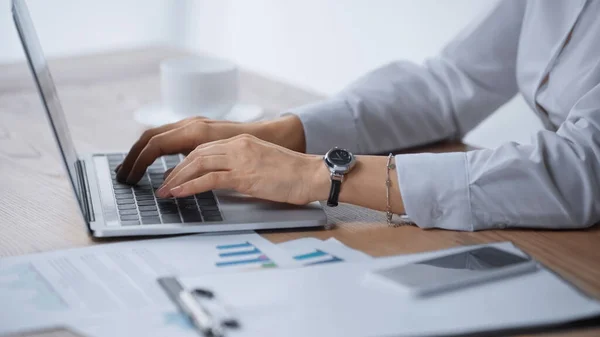 Vue recadrée de la femme tapant sur ordinateur portable près des infographies sur le bureau — Photo de stock