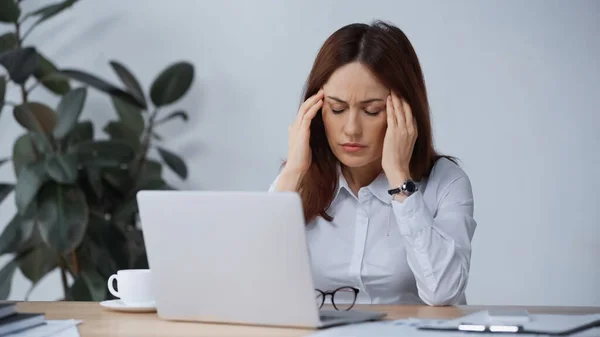 Mujer de negocios con los ojos cerrados tocando la cabeza mientras sufre de migraña en el lugar de trabajo — Stock Photo