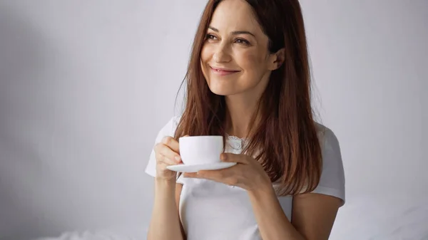 Mujer alegre sosteniendo la taza de café mientras mira hacia otro lado sobre fondo gris - foto de stock