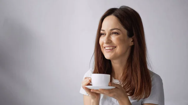 Mujer alegre mirando hacia otro lado mientras sostiene la taza de café blanco sobre fondo gris - foto de stock