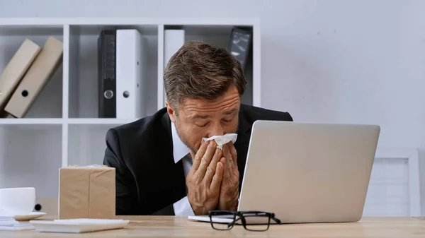 Empresário doente com olhos fechados espirrando em guardanapo de papel perto do laptop — Fotografia de Stock