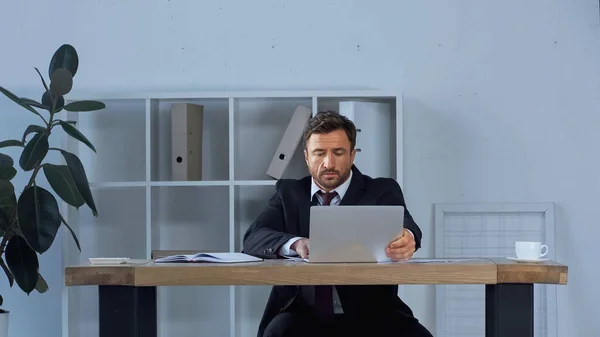 Businessman in black suit working at laptop near coffee cup on office desk — Stock Photo