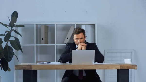 Pensativo hombre de negocios en traje sentado con las manos apretadas cerca de la computadora portátil en la oficina - foto de stock