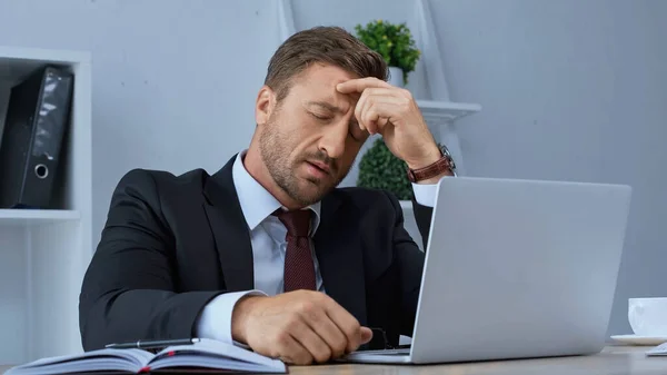 Hombre de negocios cansado que sufre de dolor de cabeza mientras está sentado en el lugar de trabajo cerca del ordenador portátil - foto de stock