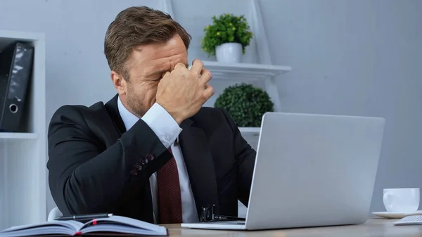 Exhausted businessman rubbing eyes while suffering from migraine near laptop — Stock Photo