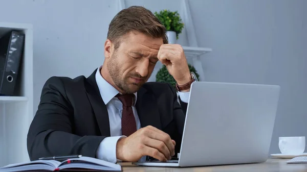 Exhausted businessman sitting with closed eyes near laptop and suffering from headache — Stock Photo