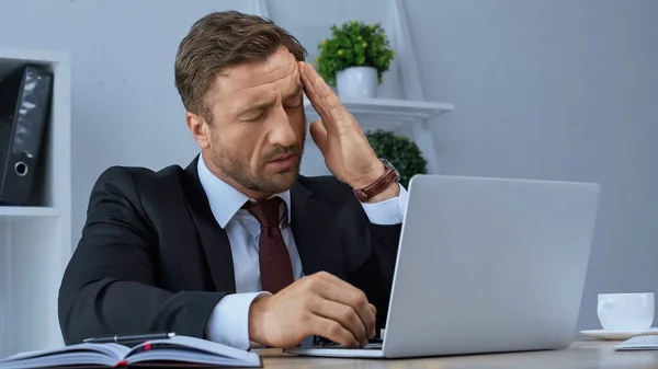 Втомлений бізнесмен сидить біля ноутбука з закритими очима і страждає від головного болю — стокове фото