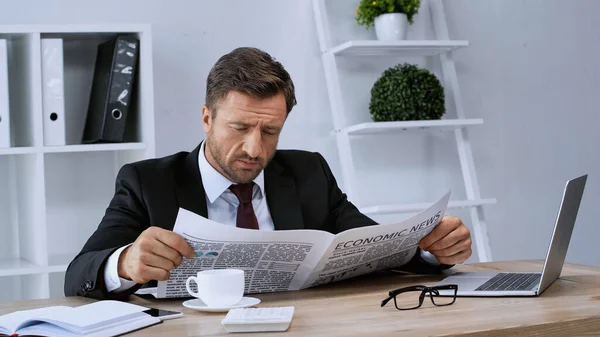 Бизнесмен в черном костюме читает экономические новости возле чашки кофе и ноутбука — стоковое фото