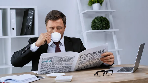 Uomo d'affari che beve caffè e legge giornali vicino al computer portatile in ufficio — Foto stock