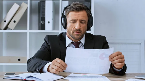 Бізнесмен в навушниках, що працюють з документами біля смартфона і ноутбука на столі — стокове фото