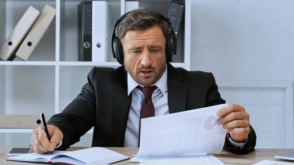 Hombre de negocios en auriculares escribiendo en cuaderno mientras trabaja con infografías en la oficina - foto de stock