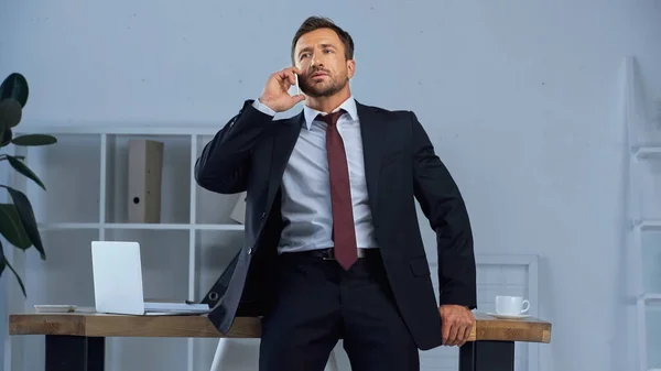 Человек в костюме разговаривает на смартфоне рядом с рабочим местом в офисе — стоковое фото