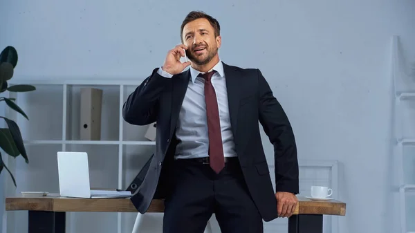 Happy man in suit talking on mobile phone near laptop and coffee cup on office desk — Stock Photo