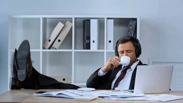 Hombre de negocios en auriculares bebiendo café mientras está sentado con las piernas en el escritorio cerca de la computadora portátil - foto de stock