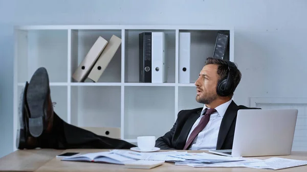 Geschäftsmann mit Kopfhörer sitzt mit Beinen auf dem Schreibtisch, während er in der Kaffeepause Musik hört — Stockfoto