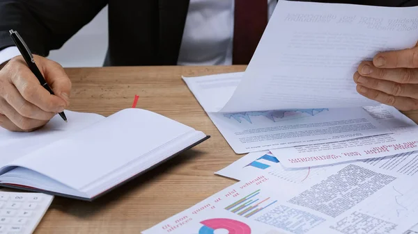 Vista recortada del empresario escribiendo en cuaderno cerca de papeles con gráficos en el escritorio — Stock Photo