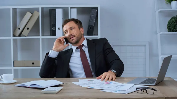 Businessman in suit talking on mobile phone near infographics and laptop on desk — Stock Photo