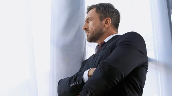 Vista de ángulo bajo del hombre de negocios en traje negro de pie con los brazos cruzados cerca de la ventana - foto de stock