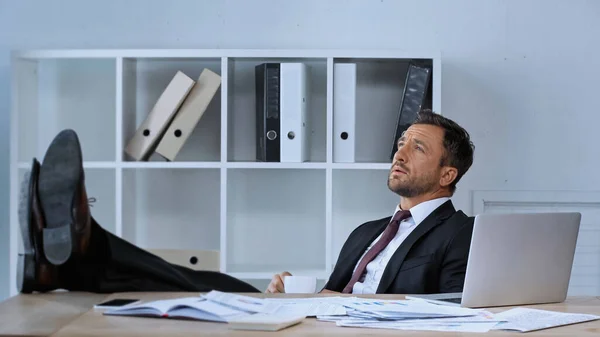 Uomo in giacca e cravatta seduto con le gambe sulla scrivania mentre riposa durante la pausa caffè in ufficio — Foto stock