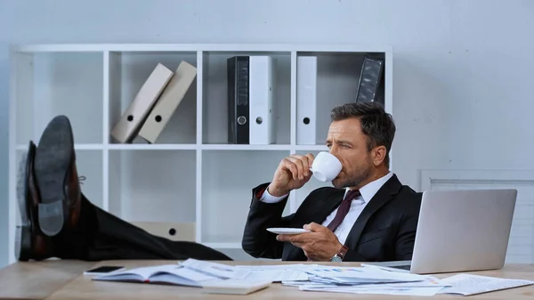 Empresário sentado com as pernas na mesa durante coffee break no escritório — Fotografia de Stock