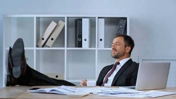 Homme d'affaires heureux assis avec les jambes sur le bureau tout en se reposant pendant la pause café — Photo de stock