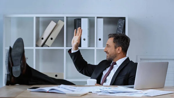 Vue latérale de l'homme heureux agitant la main tout en étant assis avec les jambes sur le bureau au travail — Photo de stock