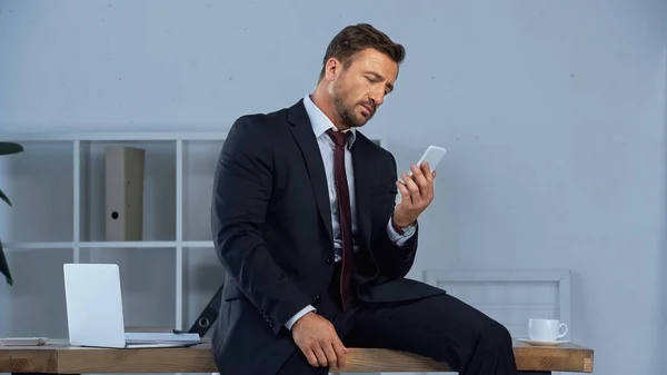 Businessman with smartphone sitting on desk near laptop and coffee cup — Stock Photo