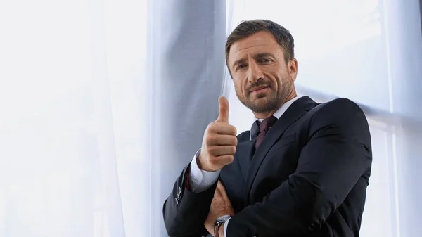 Low angle view of successful businessman showing thumb up near window in office — Stock Photo