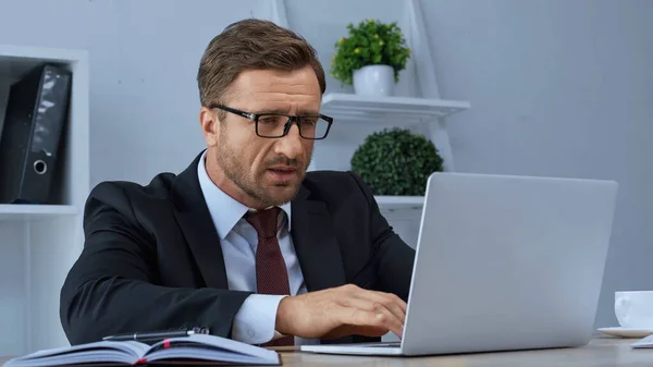 Homem de negócios em óculos digitando no laptop perto de notebook na mesa no escritório — Fotografia de Stock