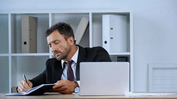 Бизнесмен, работающий возле ноутбука и пишущий в блокноте в офисе — стоковое фото