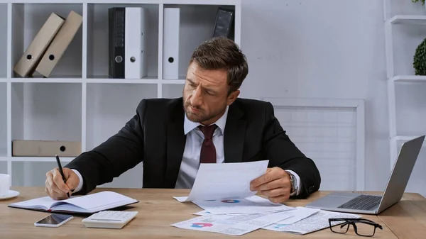 Geschäftsmann schreibt in Notizbuch, während er mit Papieren in Laptopnähe arbeitet — Stockfoto