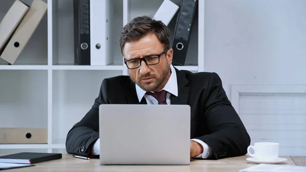 Hombre de negocios enfocado en anteojos que trabajan en la computadora portátil en la oficina - foto de stock