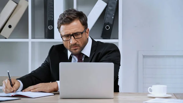 Businessman in eyeglasses writing in notebook while looking at laptop in office — Stock Photo