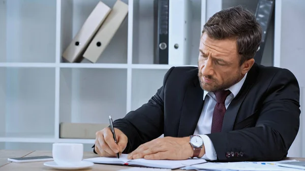 Businessman writing in notebook near coffee cup on work desk — Stock Photo