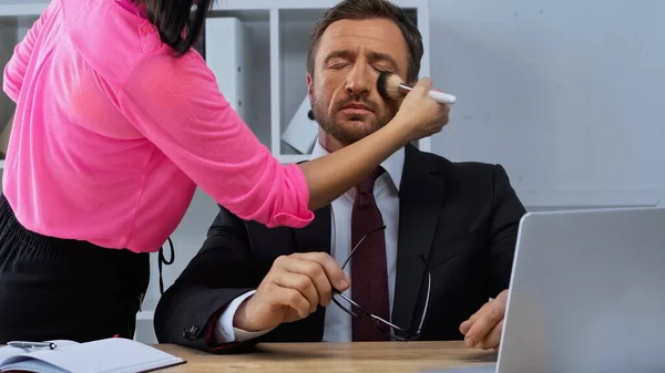 Femme faisant maquillage à l'homme en costume assis au bureau près d'un ordinateur portable — Photo de stock