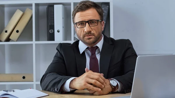 Hombre de negocios en traje negro y anteojos mirando a la cámara cerca de la computadora portátil - foto de stock