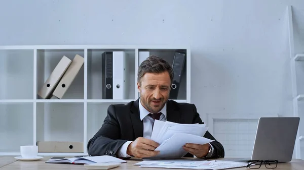 Усміхнений бізнесмен, що працює з документами біля ноутбука в офісі — стокове фото