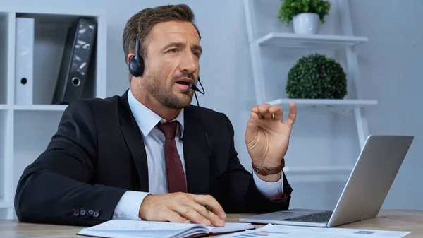 Hombre en auriculares hablando y haciendo gestos cerca de la computadora portátil en el lugar de trabajo - foto de stock