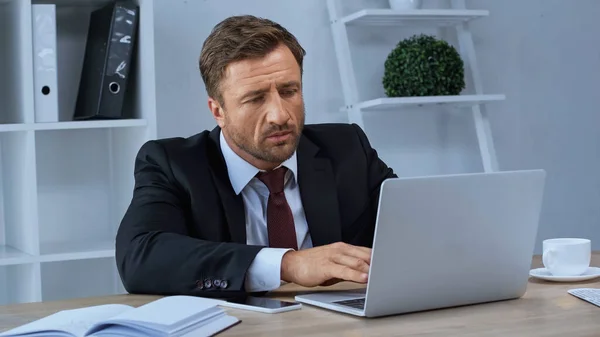 Geschäftsmann tippt auf Laptop neben Smartphone und Notebook auf Schreibtisch — Stockfoto