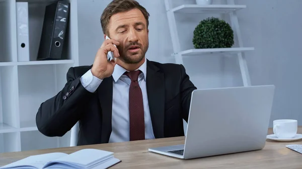 Mann in offizieller Kleidung telefoniert im Büro mit Smartphone in der Nähe von Laptop und Notebook — Stockfoto