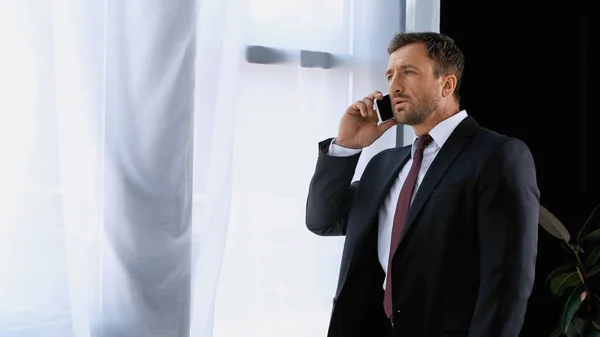 Бизнесмен в костюме стоит у окна офиса и разговаривает по мобильному телефону — стоковое фото