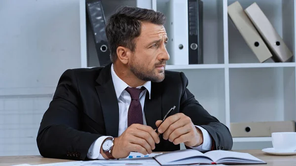 Hombre de traje negro mirando hacia otro lado mientras está sentado en el lugar de trabajo cerca de papeles y cuaderno - foto de stock
