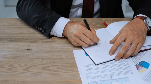 Partial view of businessman writing in notebook near infographics on work desk — Stock Photo