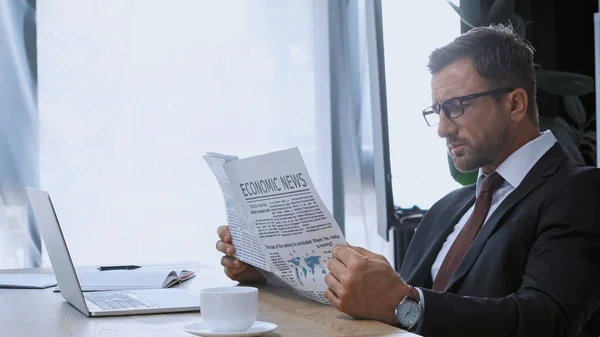 Uomo d'affari in occhiali che legge notizie economiche vicino a tazza di caffè e computer portatile — Foto stock