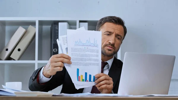 Geschäftsmann hält Dokumente mit Grafiken bei Videoanruf auf Laptop — Stockfoto
