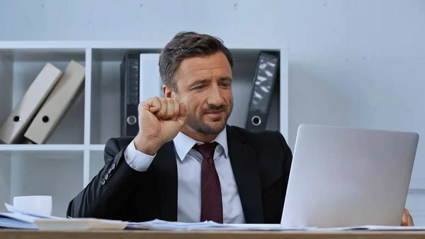 Улыбающийся бизнесмен, показывающий победный жест во время видеочата на ноутбуке — стоковое фото